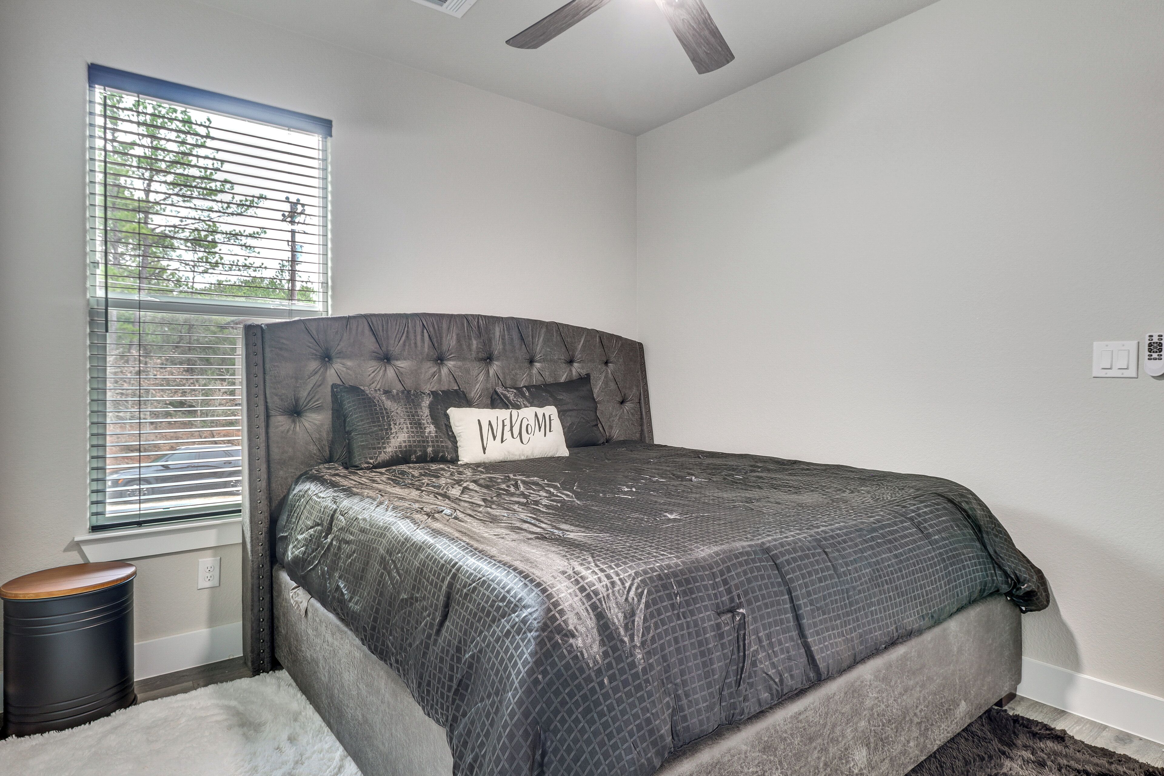 A bedroom with a large bed, a window with blinds, a ceiling fan, and a small trash can.