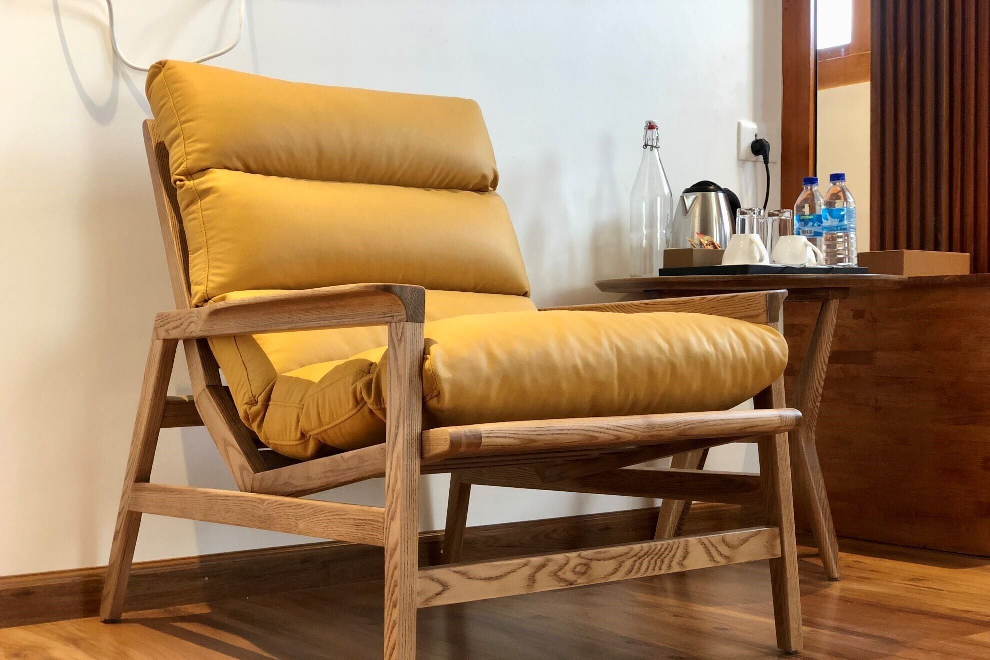 Una silla de madera con cojines amarillos en una habitación con piso de madera y una mesita con una tetera, tazas y botellas.