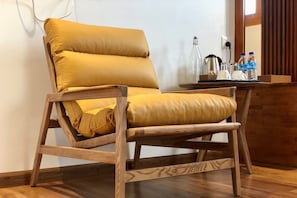 Une chaise en bois avec des coussins jaunes, dans une pièce dont le sol est en bois, et où se trouve une petite table avec une bouilloire, des tasses et des bouteilles.