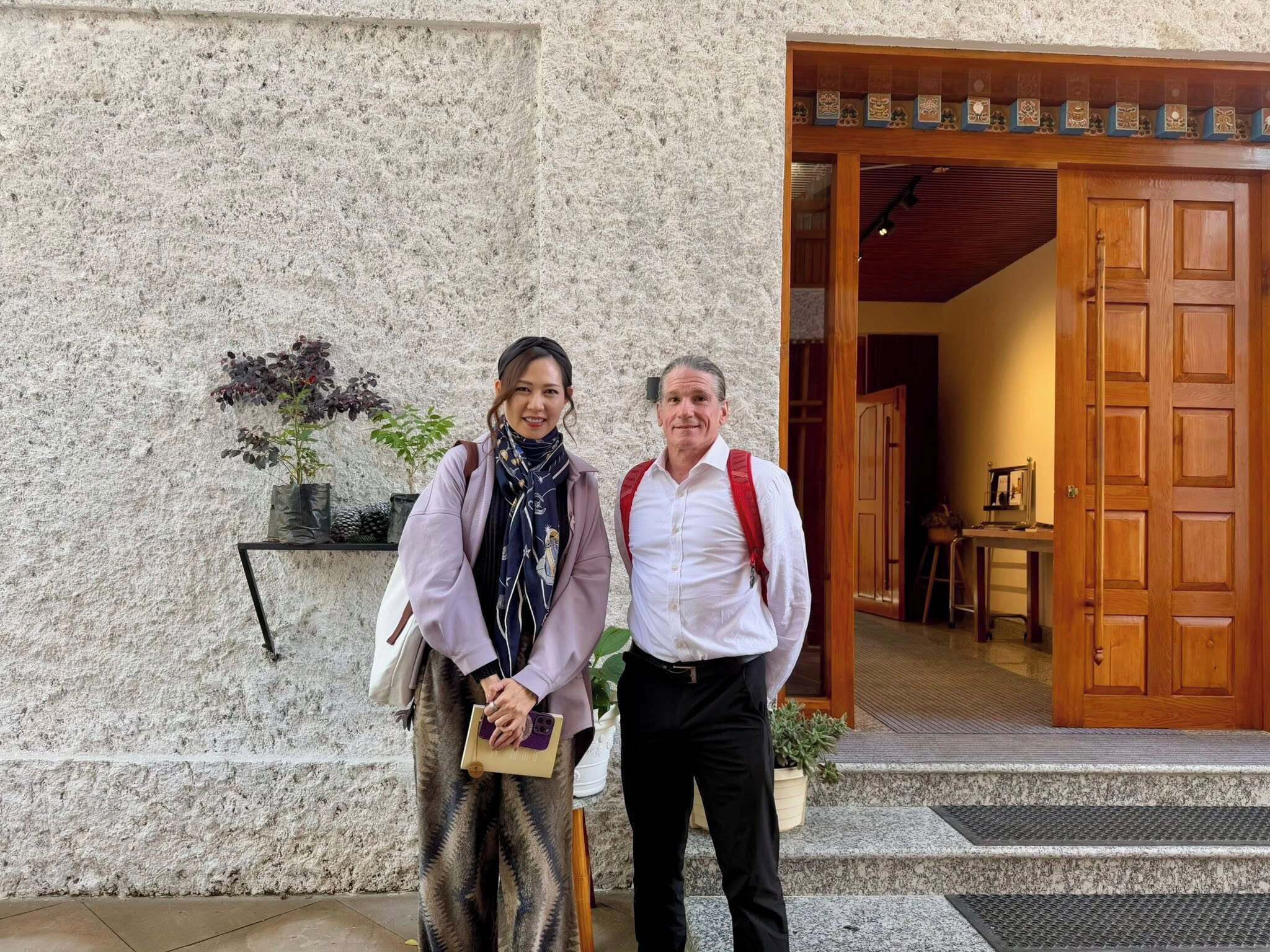 Two people standing in front of a building with a stone wall and a wooden door.