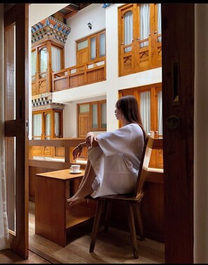 Eine Person sitzt an einem Holztisch in einem Raum mit hoher Decke und Holzbalken, mit Blick auf einen Balkon mit Holzgeländern und einer gemusterten Decke.