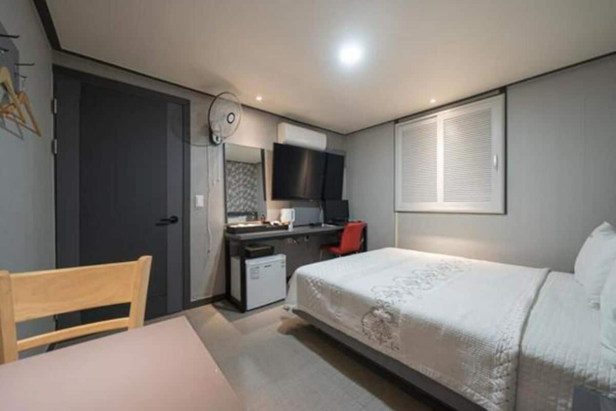 A compact hotel room with a single bed, a desk with a chair, a small kitchenette, and a window with blinds.