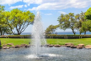 A fountain with water jets in a landscaped area with trees and a view of the sea. - Kamaole Sands #10-313 Ocean View, Large Lanai, Walk to the Beach (Kihei)