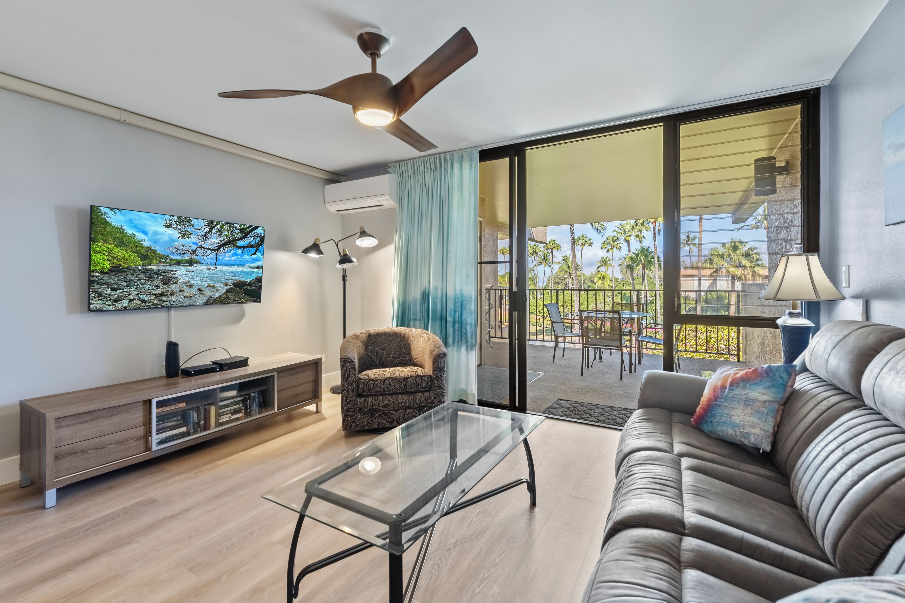 A modern living room with a flat-screen TV, a glass coffee table, a leather sofa, a patterned armchair, and a ceiling fan.
