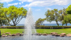 Eine Fontäne mit Wasserstrahlen in einer angelegten Grünanlage mit Bäumen und Blick aufs Meer.