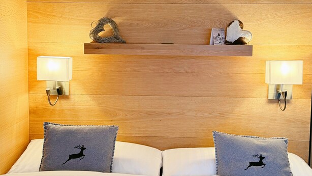 A hotel room with a wooden headboard, two bedside tables with wall-mounted lamps, and two beds with matching pillows. - Chalet Achat by Zermatt Premium Apartments (Zermatt)