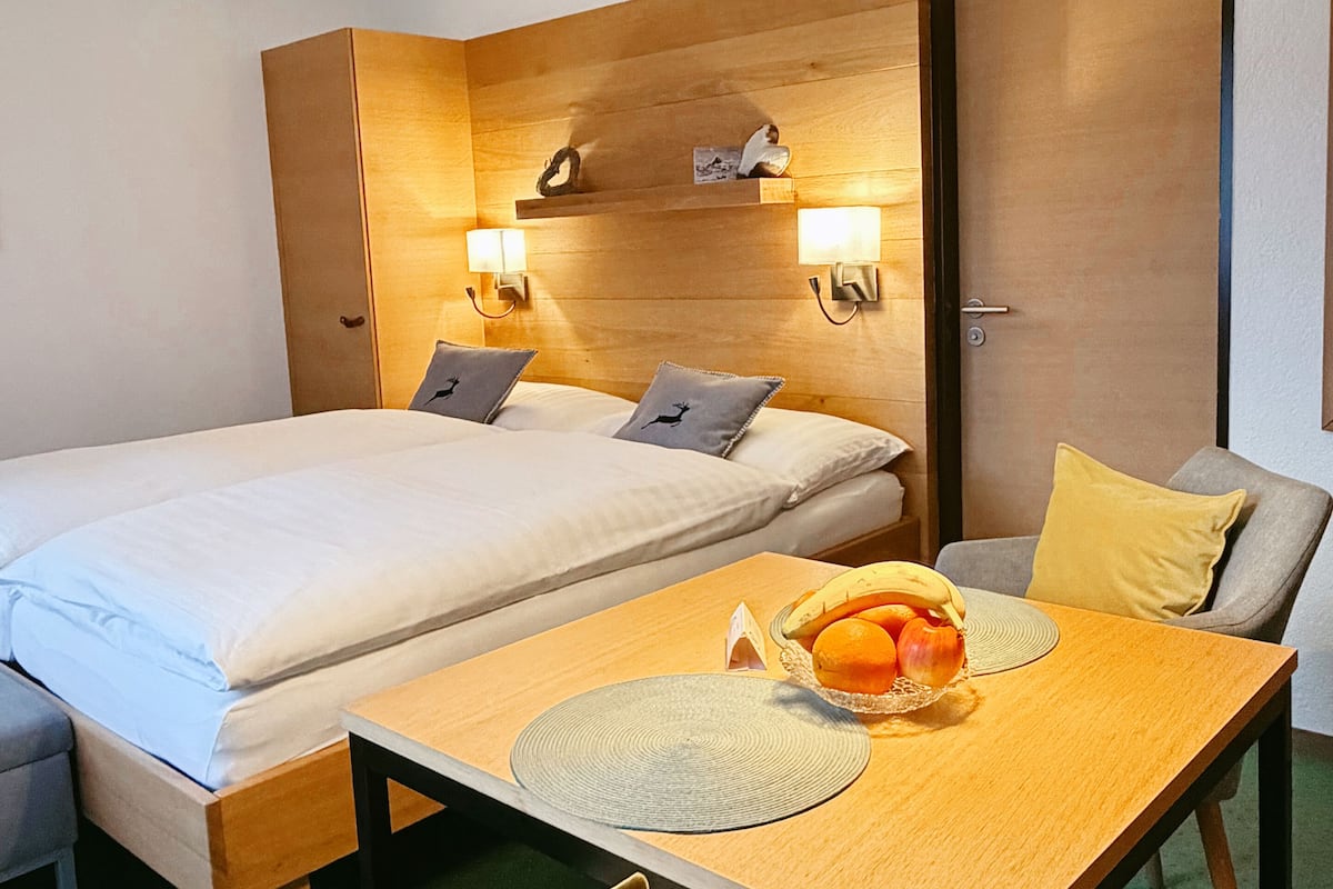A hotel room with a bed, bedside tables, a dining table with a fruit bowl, and a chair.