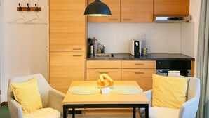 A modern hotel room with a bed, two armchairs, a small table, a kitchen area with wooden cabinets, a black pendant light, and a window with curtains. - Chalet Achat by Zermatt Premium Apartments (Zermatt)
