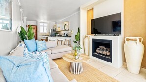 A modern living room with a sofa, coffee table, and fireplace. There is a kitchen in the background with bar stools. - Atlantic Terraces 19 (Cape Town)