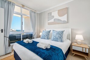 Una habitación de hotel con una cama grande, una ventana con cortinas, una mesita de noche con lámpara y un cuadro abstracto colgado en la pared.