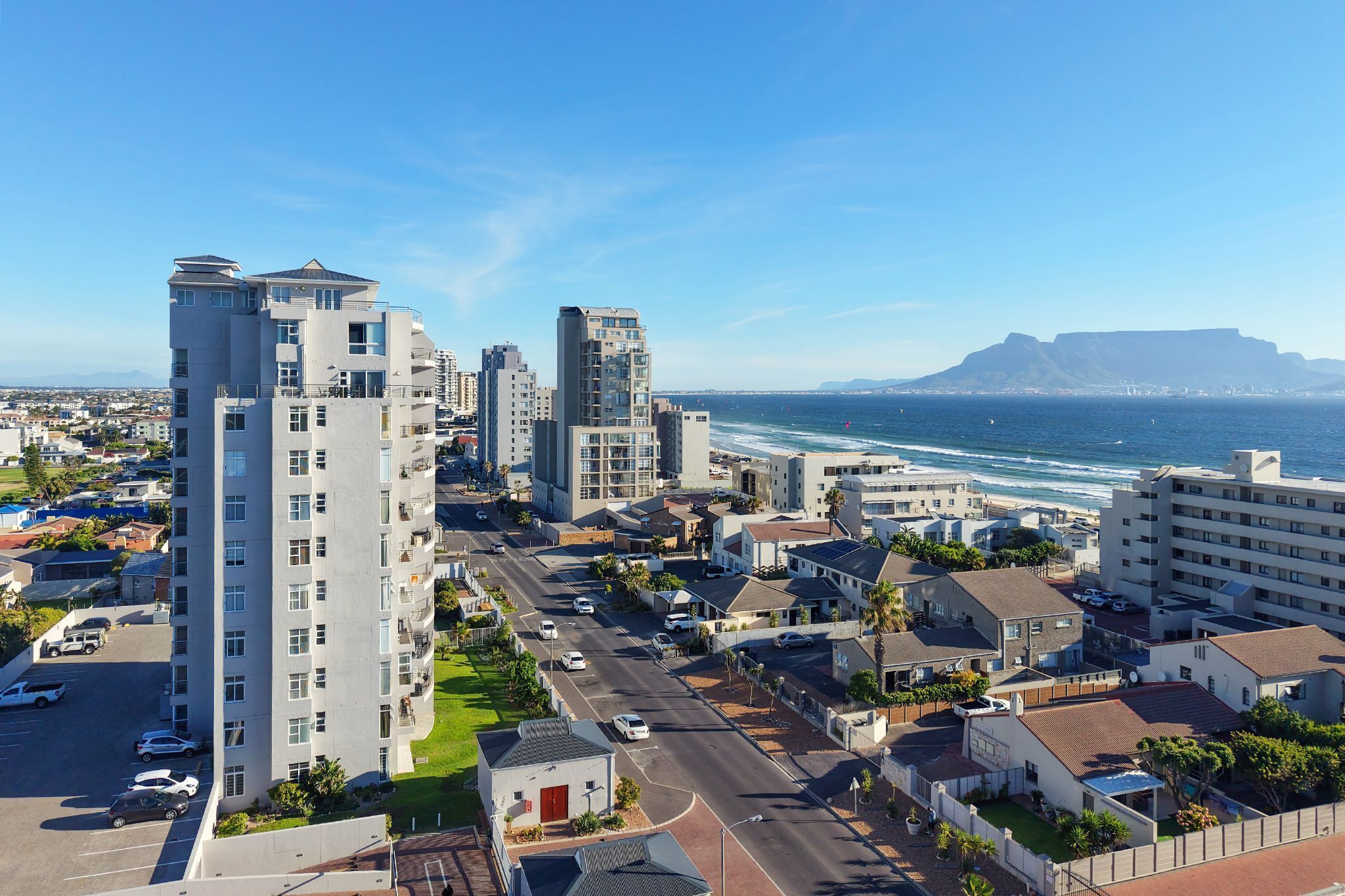 Uma área urbana costeira com edifícios altos, uma estrada com carros e vista para o oceano e uma montanha ao longe.