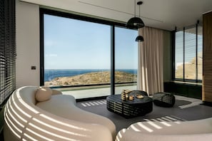 A modern bedroom with a large sliding glass door, a curved sofa, a round coffee table, and a view of a coastal landscape. - New Luxury Villa w/ Private Pool, Spa & Sea Views! (Heraklion)