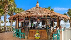 Eine Tiki-Bar mit einem Reetdach, einer Holztresen und Menütafeln. Türkisfarbene Stühle und eine Ananasdekoration sind vorhanden.