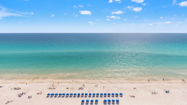 Ein Strand mit klarem, blauem Wasser, einem Sandufer und einer Reihe blauer Sonnenschirme.