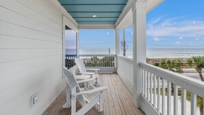 Eine überdachte Veranda mit weißen Geländern und weißen Stühlen, mit Blick auf einen Strand unter blauem Himmel.