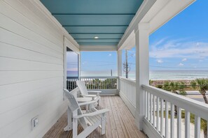Une véranda couverte, dotée de balustrades blanches et de chaises blanches, qui offre une vue sur une plage sous un ciel dégagé.