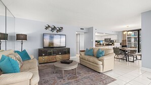 A spacious living room with a beige sofa, a coffee table, and a television. There is a kitchen in the background with a dining area. - Buoy Oh Buoy! Sun, Sand, and Smiles Await! Sleeps 8! (Panama City Beach)