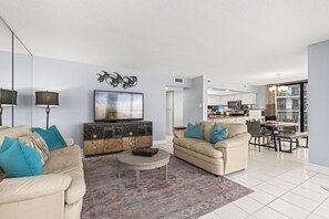Ein geräumiges Wohnzimmer mit einem beigen Sofa, einem Couchtisch und einem Fernseher. Im Hintergrund ist eine Küche mit Essbereich zu sehen.