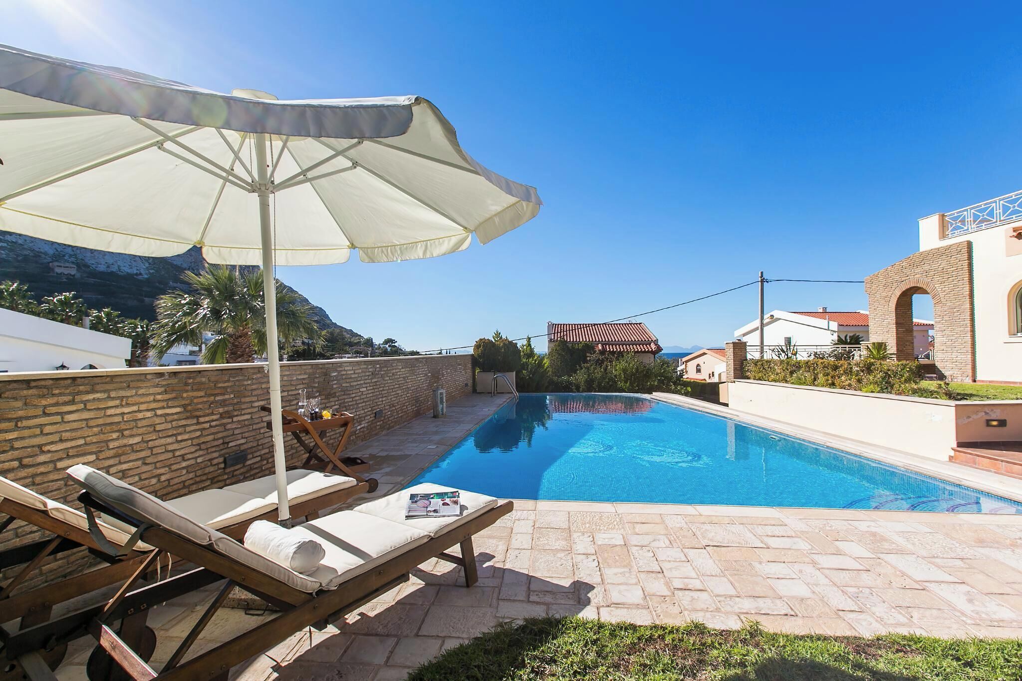 A swimming pool with lounge chairs and an umbrella, surrounded by a brick wall and a mountain view.