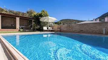 A clear blue swimming pool with lounge chairs and an umbrella, surrounded by a brick wall and a hillside in the background.