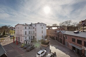 Un edificio de varios pisos con fachada blanca, un estacionamiento con varios coches y un edificio de ladrillo con techo metálico al fondo.