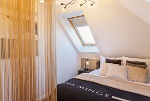 Un dormitorio con techo inclinado, tragaluces, cama con muchos almohadones, mesita de noche con lámpara y pared con listones de madera verticales.