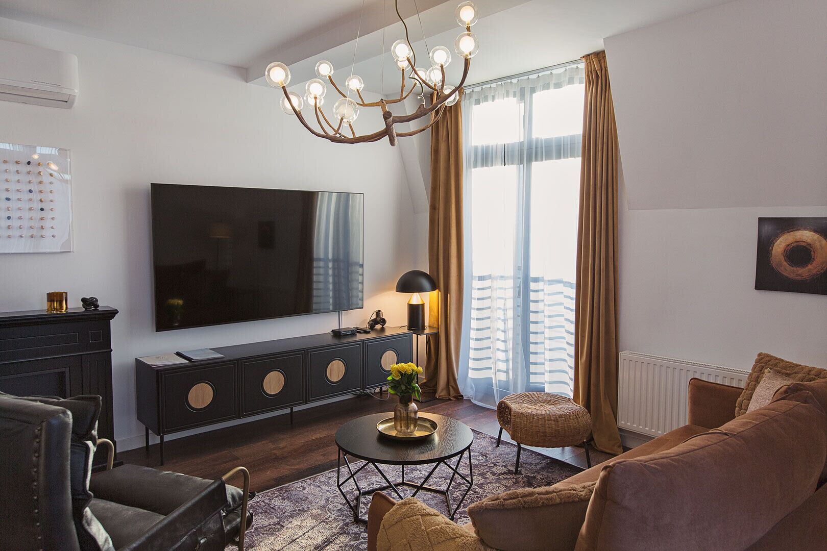 A modern living room with a flat-screen TV, dark wood furniture, a chandelier, and a large window with curtains.