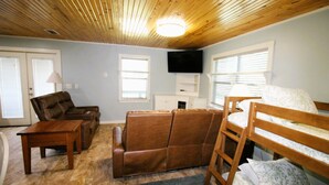 Una habitación con techo de madera, un sofá de cuero marrón, un banco de madera, una mesita de madera, un televisor montado en la pared y una ventana con persianas.