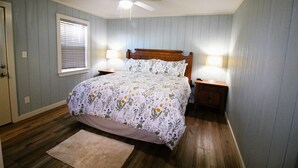 Un dormitorio con una cama de madera, dos lámparas de noche, un ventilador de techo, una ventana con persianas y piso de madera.