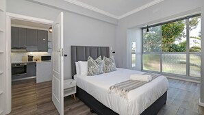 A bedroom with a large bed, a nightstand, a built-in desk, and a view of the outdoors through a large window. - Modern Two Bed Central Green Point Stadium (Cape Town)