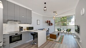 A modern kitchen with grey cabinets, a built-in oven, and a central island with bar stools. There is a dining area with a table and chairs, and a large window providing natural light. - Modern Two Bed Central Green Point Stadium (Cape Town)