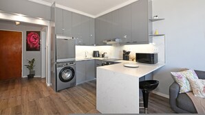 A compact kitchen with grey cabinets, a built-in washing machine, and a small island with a microwave. - Modern Two Bed Central Green Point Stadium (Cape Town)