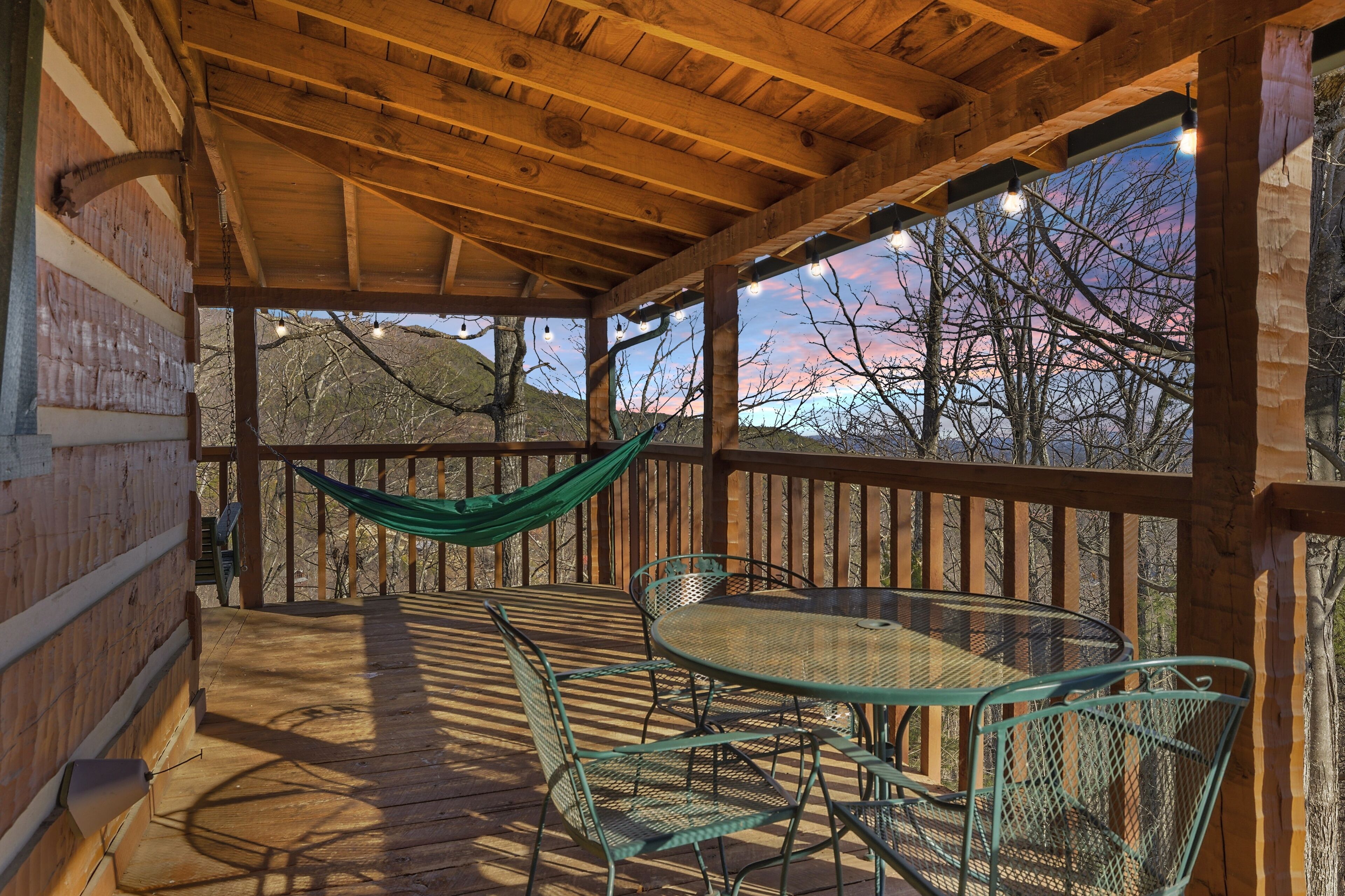 A wooden deck with a table and chairs, a hammock, and a view of trees and hills.