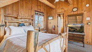 Un dormitorio de cabaña de madera con una cama, una mesita de noche, un aparador, un ventilador de techo y un cuadro de un paisaje.
