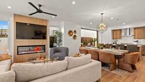 A modern living room with a sofa, a coffee table, and a TV mounted on a wooden wall. There is a dining area with a table and chairs, and a kitchen with wooden cabinets and a countertop. - Simply Paradise 5 Bedroom Townhouse (St. George)