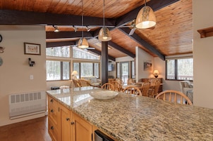 A spacious kitchen with granite countertops, wooden cabinets, and a large island. There are multiple hanging pendant lights and a ceiling fan.