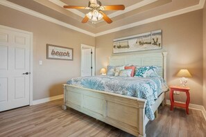 A bedroom with a bed, a ceiling fan, a nightstand, a lamp, and a wall with a 'beach' sign. - Sea Worthy 3 Bedroom Home (Port Bolivar)