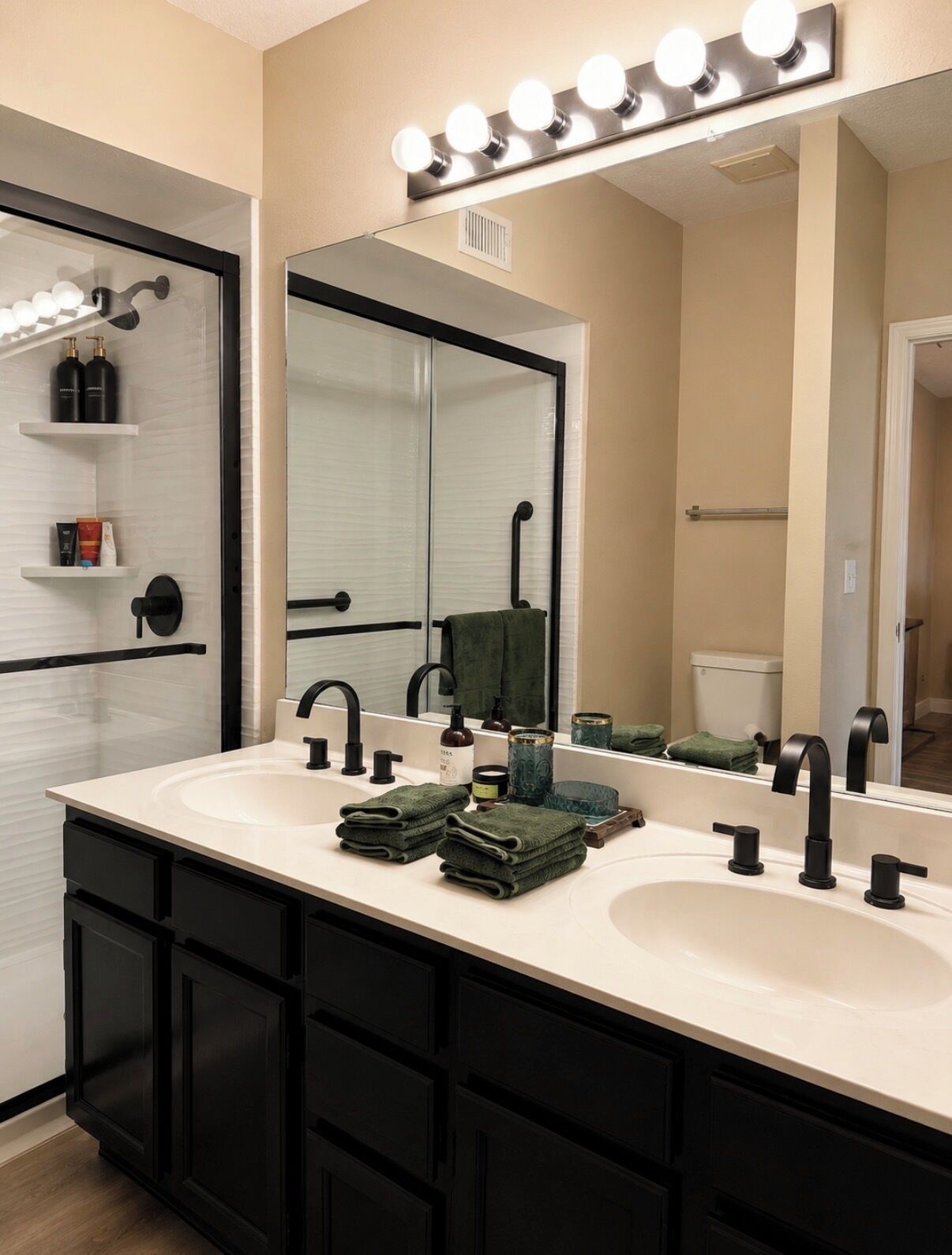 A bathroom with a double sink, black cabinets, a large mirror with lights, and a towel rack.