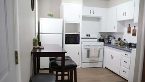A compact kitchen with white cabinets, a microwave, an oven, and a sink. There are two stools and a small table.