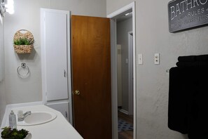 Un baño con un lavamanos blanco, una puerta de madera, una cesta con plantas y un cartel en la pared.