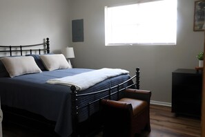 Una cama bien hecha con una colcha azul oscuro y almohadas blancas, una mesita de noche con una lámpara, y una ventana que deja entrar luz natural.