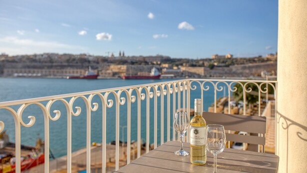 Une table avec une bouteille de vin et deux verres, donnant sur une étendue d’eau où l’on aperçoit des bateaux et, au loin, le paysage urbain.