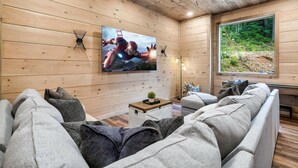 A cozy cabin interior with a wooden ceiling, a large window with a forest view, a comfortable couch, and a wall-mounted TV displaying a superhero action scene. - Kids LOVE This Cabin! Racing Games + Hot Tub (Sevierville)