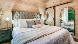 A bedroom with a wooden ceiling, a large bed with a tufted headboard, a nightstand with a lamp, and a mirror reflecting a window view. - Kids LOVE This Cabin! Racing Games + Hot Tub (Sevierville)