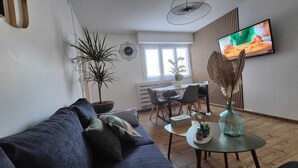 A modern living room with a dark blue sofa, a dining table with chairs, a TV mounted on the wall, and a potted plant. - Between lakes and mountains (Cluses)