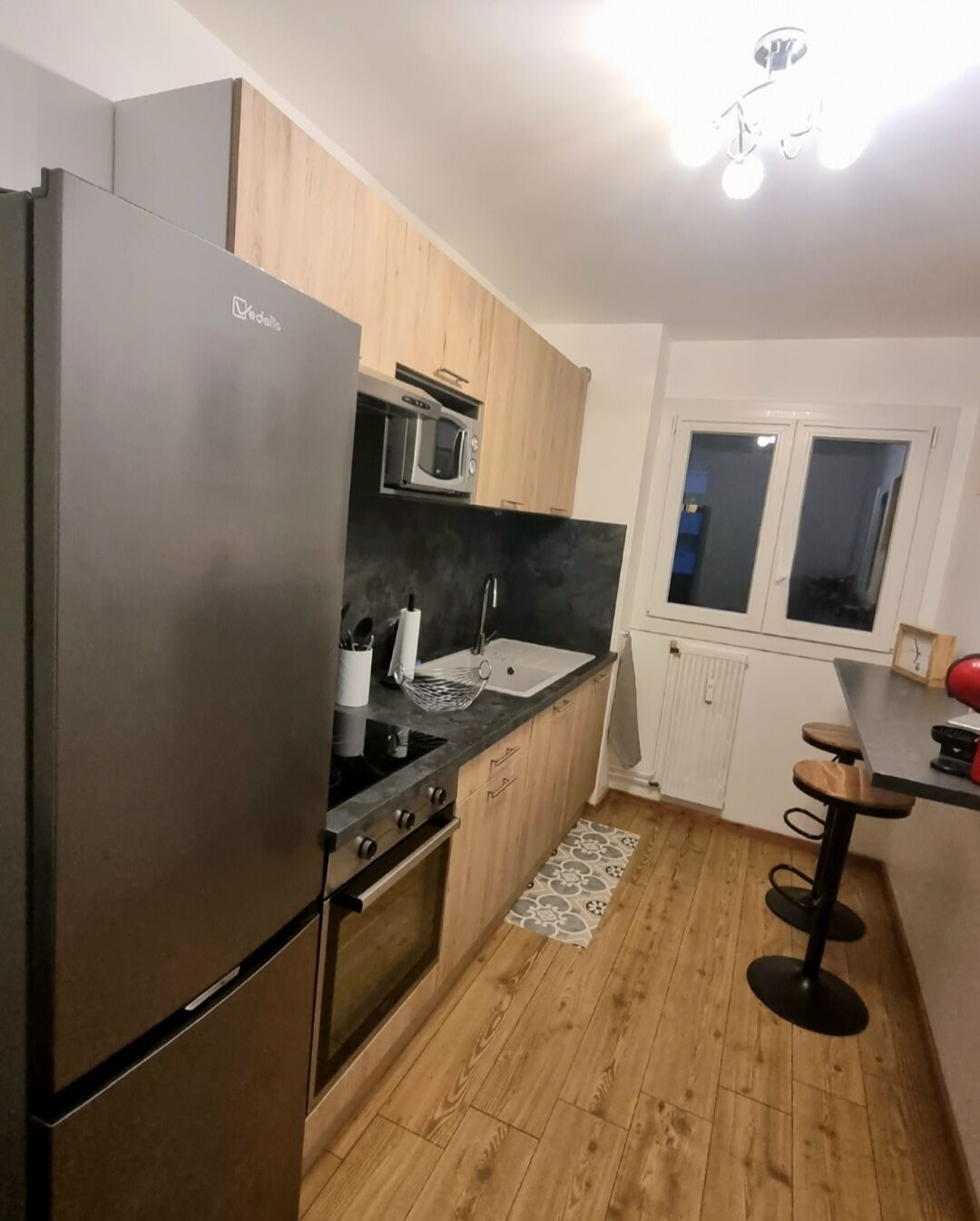 A compact kitchen with a refrigerator, microwave, and stove. It features wooden cabinets, a black countertop, and a small window.