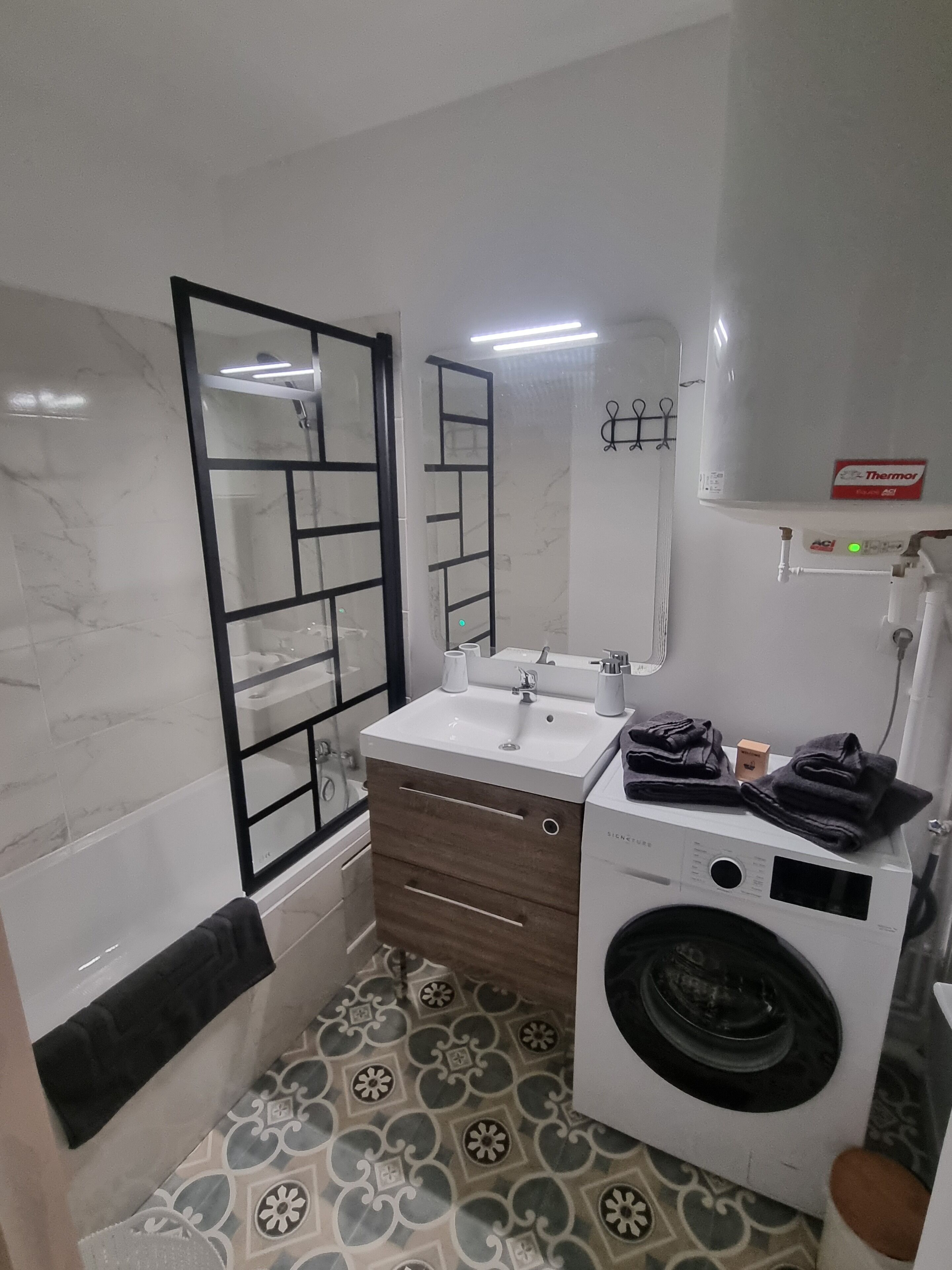 A modern bathroom with a white sink, wooden vanity, a glass shower enclosure, a washing machine, and patterned floor tiles.