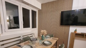 A dining area with a wooden table set for four, green chairs, a TV mounted on the wall, and a window with blinds. - Between lakes and mountains (Cluses)