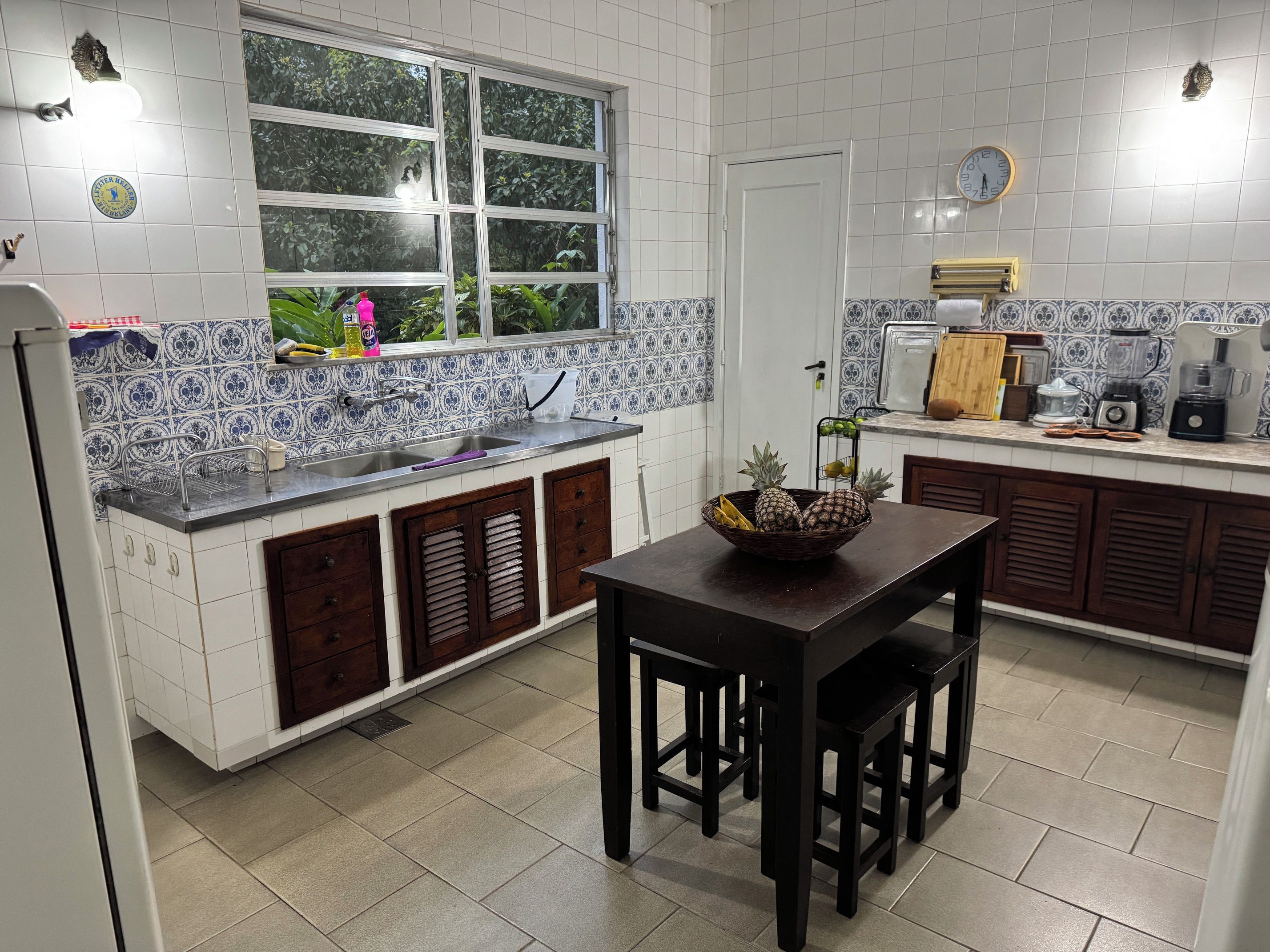 Uma cozinha com janela, pia, mesa com cesta de frutas e um relógio na parede.
