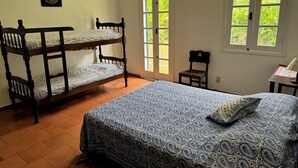 Une chambre avec un lit superposé, un lit simple avec une couvre-lit à motifs, une chaise et une fenêtre donnant sur un paysage verdoyant.